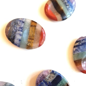 Op de afbeelding: Verschillende gepolijste, ovaalvormige stenen met een gestreept patroon van verschillende kleuren, waaronder blauw, rood, geel en bruin. De stenen zijn gerangschikt op een wit oppervlak, waardoor hun natuurlijke texturen en tinten worden benadrukt. Deze stenen worden waarschijnlijk gebruikt voor decoratieve of spirituele doeleinden.