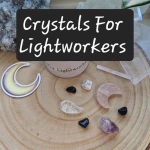 May include: A collection of crystals and a moon-shaped sticker arranged on a wooden surface. The crystals include black, clear, and purple stones, along with a pink crescent moon-shaped crystal. A container with the text "Crystals For Lightworkers" is also present.