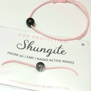 May include: A pink string bracelet with a black shungite bead. The bracelet is on a white card with the text "For Protection Shungite From 5G/ EMF/ Radio Active Waves www.crystalwaterslove.etsy.com"