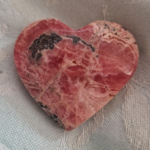 May include: A heart-shaped stone with a pink and red marbled pattern. The stone has dark gray and black inclusions. The heart is resting on a light blue fabric.