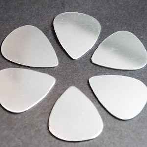 May include: Six silver guitar picks, shaped like a teardrop, arranged in a circle on a gray background.