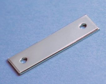 Puede incluir: Una placa rectangular de metal plateado con dos agujeros redondos. La placa tiene una superficie lisa y pulida.
