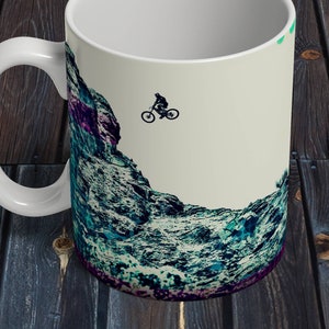 May include: White ceramic mug with a graphic of a mountain range and a silhouette of a person riding a mountain bike. The background is a light beige color.