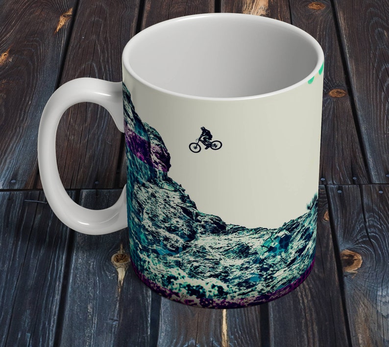 May include: White ceramic mug with a graphic of a mountain range and a silhouette of a person riding a mountain bike. The background is a light beige color.