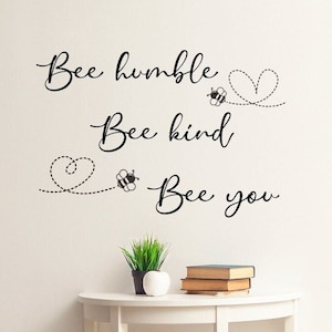 May include: White wall with black lettering that reads "Bee humble, Bee kind, Bee you". Three black and yellow bees are flying with hearts behind them.