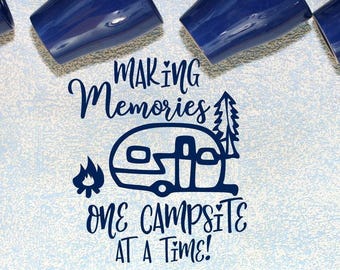 May include: A dark blue vinyl decal featuring a camper trailer, a campfire, and a pine tree. The text reads "MAKING MEMORIES ONE CAMPSITE AT A TIME!" with small heart accents. The design is displayed against a light blue textured background.
