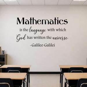 Könnte beinhalten: Eine Klassenraumwand mit einem Zitat in schwarzer Schrift: „Mathematik ist die Sprache, mit der Gott das Universum geschrieben hat.“ Das Zitat wird Galileo Galilei zugeschrieben. Im Vordergrund sind Schreibtische und Stühle zu sehen.