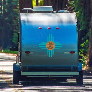 May include: A silver camper trailer with a blue and turquoise Zia symbol on the back. The camper is parked on a paved road with trees on either side.