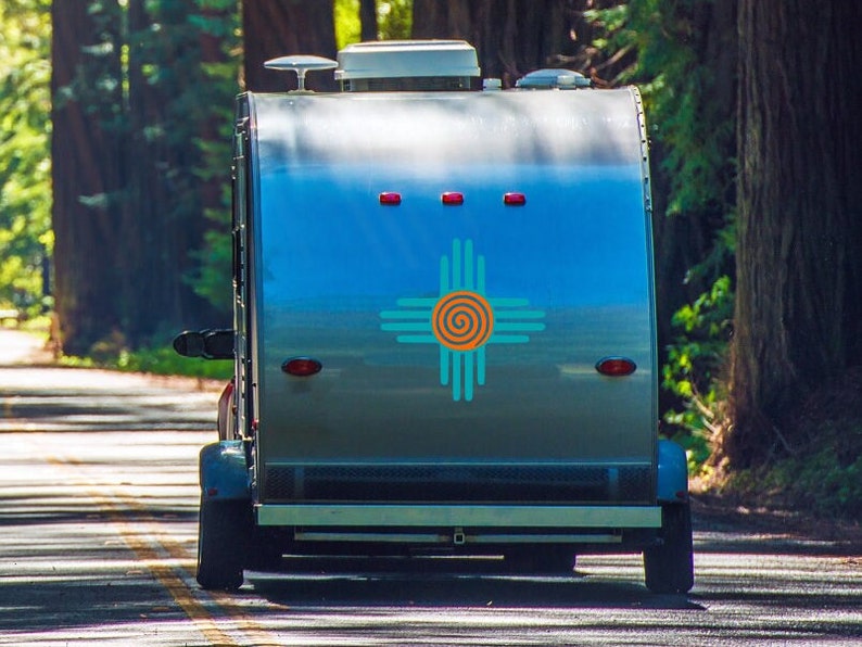 May include: A silver camper trailer with a blue and turquoise Zia symbol on the back. The camper is parked on a paved road with trees on either side.