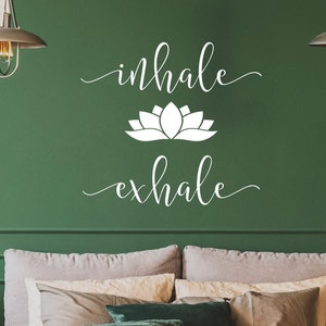 May include: White vinyl wall decal with the words "inhale" and "exhale" in a cursive font. A white lotus flower graphic is between the words. The decal is on a green wall above a bed with white and beige pillows.