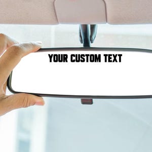 May include: A rectangular vehicle rearview mirror featuring a black frame and a white reflective surface. The text YOUR CUSTOM TEXT is printed in bold black capital letters across the upper center portion of the mirror's reflective surface.
