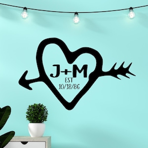 May include: Black heart-shaped wall decal with an arrow through it. The initials "J+M" are inside the heart, with "EST 10/18/86" below. The decal is on a light blue wall, with string lights above.
