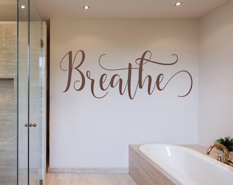Puede incluir: Un adhesivo de pared con la palabra Breathe escrita en una fuente cursiva fluida de color marrón. Las letras están aplicadas directamente sobre una superficie de pared lisa y clara, situadas sobre una bañera blanca en un baño.