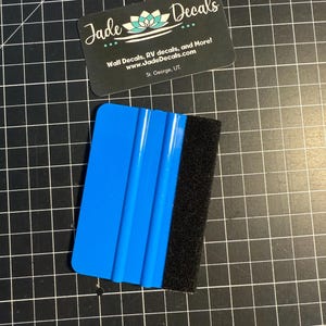 May include: A blue plastic squeegee tool with a black felt edge, accompanied by a black rectangular business card featuring the text Jade Decals, Wall Decals, RV decals, and More!, www.JadeDecals.com, and St. George, UT.