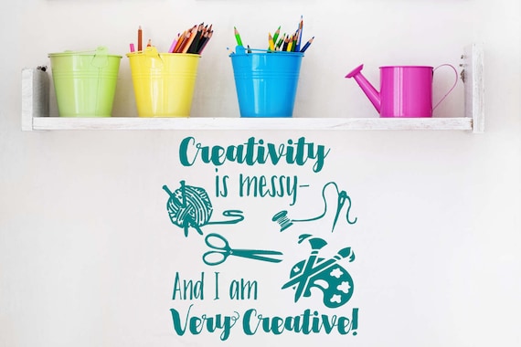 A white shelf hung on a white wall. The shelf holds three buckets in green, yellow and blue that hold colored pencils. There is a purple watering can on the right side of the shelf. Under the shelf is decal that has sea-green text and illustrations of yarn, thread, a needle and an art pallet with the words "Creativity is messy -and I am very creative!" Art posters and wall decals make excellent gifts for crafters. 