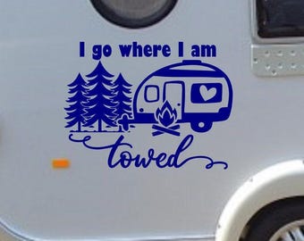 May include: A dark blue vinyl decal featuring a camper trailer, three pine trees, and a campfire. The text reads "I go where I am towed" in a combination of block and cursive lettering. The design is applied to a light-colored surface.