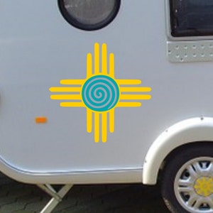 May include: A white camper van with a yellow and blue stylized symbol of a spiral inside a circle with four arms extending out from the circle. The symbol is commonly known as the "Sun Symbol" or "Southwest Symbol".