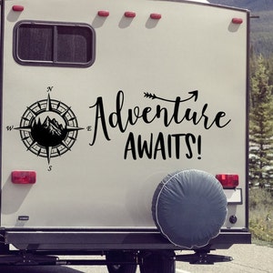 May include: A decal on the back of a camper with the words "Adventure Awaits!" in black script. A compass rose with a mountain scene is on the left. A spare tire is covered with a gray cover.