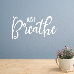 May include: A light blue wall with the text "JUST Breathe" in white cursive lettering. A wooden table with a small potted plant with pink flowers sits on the table.