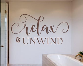 May include: A brown wall decal featuring the word relax in a flowing cursive script, positioned above the word & UNWIND in a clean, capitalised sans-serif font. The lettering is applied directly to a smooth, light-coloured interior wall surface.