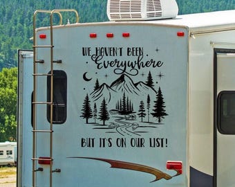 May include: A light blue recreational vehicle features a black decal with a mountain and forest landscape, a winding road, stars, and a crescent moon. The text reads: WE HAVEN'T BEEN EVERYWHERE BUT IT'S ON OUR LIST! A metal ladder is attached to the side.