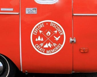 May include: A bright red camper exterior featuring a circular white decal. The decal displays crossed arrows, a campfire, pine trees, a bird, and mountains, with the text "EXPLORE DISCOVER CREATE MEMORIES" arranged around the perimeter.