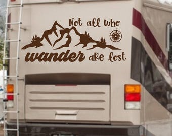 May include: A brown vinyl decal applied to the rear of a recreational vehicle. The design features a mountain range silhouette, small pine trees, a compass rose, and the text Not all who wander are lost in a stylized script font.