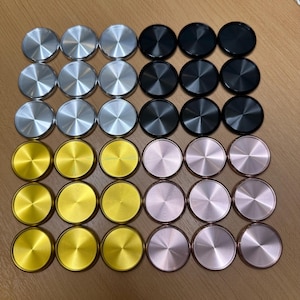 Puede incluir: Una colección de discos redondos metálicos en varios colores, incluyendo plata, negro, oro y oro rosa. Los discos están dispuestos en filas y columnas, creando un patrón visualmente atractivo.