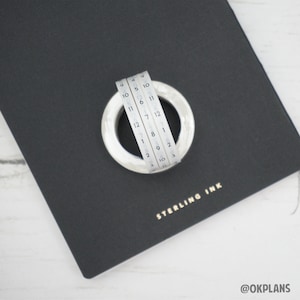 Pode incluir: Um rolo de fita washi branca com números pretos de 1 a 12, enrolado em torno de um anel branco. A fita está em um caderno preto com as palavras "STERLING INK" em letras douradas.