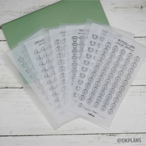 May include: A set of clear sticker sheets with black outlines of baby-related icons. The icons include a stroller, a bottle, a bib, a nappy, a first aid kit, a onesie, and a star. The text on the sheets reads "Stroller Savings", "Heal", "Tin", "Bottles + Bibs Savings Challenge", and "Total = okplans".