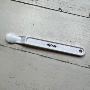 May include: White plastic spoon with a rounded end and a hole at the other end. The spoon has the word "olplans" printed on it.