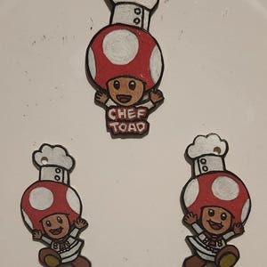 Op de afbeelding: Drie houten ornamenten van een paddenstoelfiguur met een koksmuts. Het figuur draagt een wit koksuniform en heeft een rode paddenstoelhoed met witte stippen. Het middelste ornament heeft de tekst "CHEF TOAD" erop.