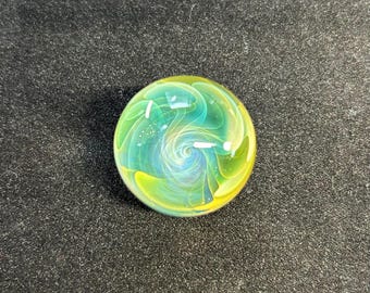 Pode incluir: Uma bola de gude de vidro redonda com um design em espiral. A bola de gude apresenta uma mistura de cores verdes, amarelas e brancas. O design tem um padrão em espiral no centro. A bola de gude está sobre um fundo cinza escuro.