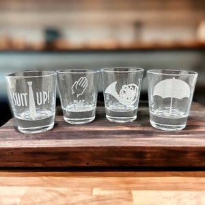 May include: Four clear glass shot glasses with different designs etched into them. The designs include a tie with the text "Suit Up!" a hand with the text "Slap the Commissioner", a French horn, and an umbrella.