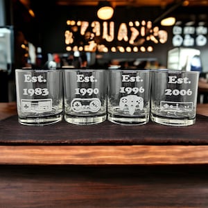 May include: Four clear glass tumblers with etched designs. Each tumbler features a different year, "Est." followed by a year, and a video game controller graphic. The years are 1983, 1990, 1996, and 2006.