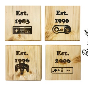 May include: Four wooden square coasters with black text and images of video game controllers. The coasters read "Est. 1983", "Est. 1990", "Est. 1996", and "Est. 2006".