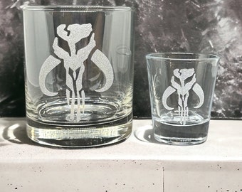 May include: Two clear glass drinking glasses with a white Mandalorian symbol etched on the front. The larger glass is a rocks glass and the smaller glass is a shot glass.