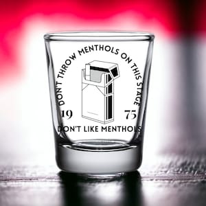 May include: A clear glass shot glass with the text "Don't Throw Menthols On This Stage 19 75 Don't Like Menthols" printed on the side. The text is in a black font and is centered on the glass. There is a black and white illustration of a pack of cigarettes on the glass.