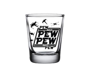 May include: Clear glass shot glass with the word "PEW" in a black, bold font, repeated three times. Black silhouettes of spaceships are above the text. The shot glass is on a white background.