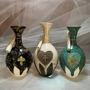 May include: Three ceramic vases with heart and cross designs. The vases are in black, cream, and teal, with gold-colored crosses. Each vase has a unique speckled pattern and a rolled paper scroll attached with copper wire.