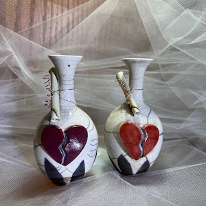 May include: Two white ceramic vases with a broken heart design. Each vase has a copper wire wrapped around the neck with a piece of paper attached.