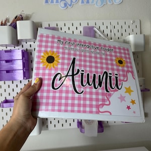 Puede incluir: Una carpeta rosa y blanca con estampado de cuadros con el texto "My first interactive folder" y "Aiuuii". La carpeta tiene gráficos de girasoles y estrellas. El fondo incluye una tabla perforada blanca y la palabra "InspiraArte".