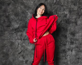 Op de afbeelding: Een rood hoodie-sweatshirt en bijpassende joggingbroek met zwarte zijstrepen. Het sweatshirt heeft een hoge hals en een trekkoord. Het model draagt witte sneakers.