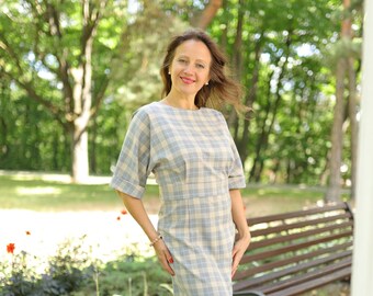 Può includere: Una donna indossa un abito a quadri blu chiaro e bianco con una silhouette aderente e maniche corte. È in piedi su un sentiero di mattoni in un parco.