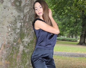 May include: A woman wearing a dark blue satin jumpsuit with a black lace trim on the shoulders. She is standing next to a tree trunk.