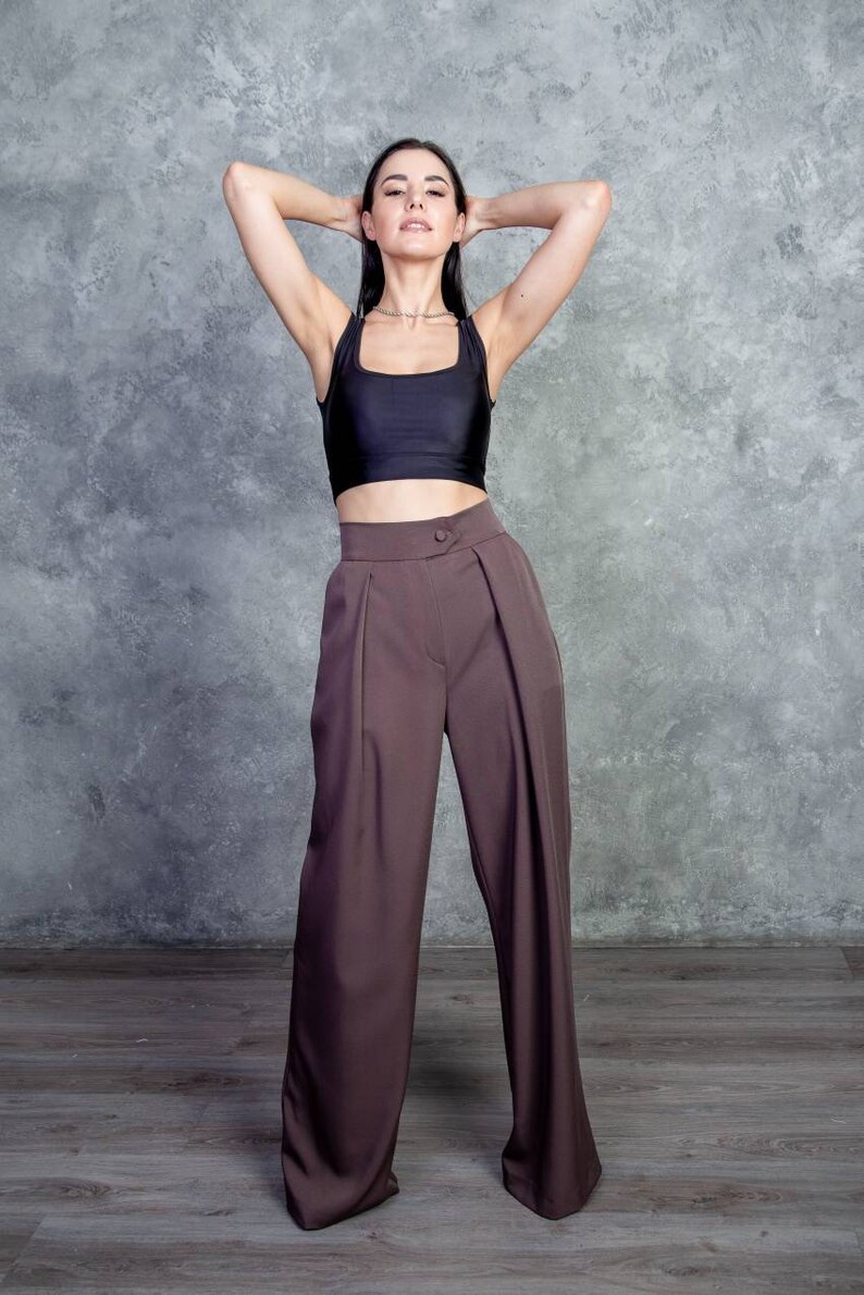 K&ouml;nnte beinhalten: Eine Frau tr&auml;gt ein schwarzes Crop-Top und eine braune weite Hose. Sie steht vor einer grauen Betonwand.