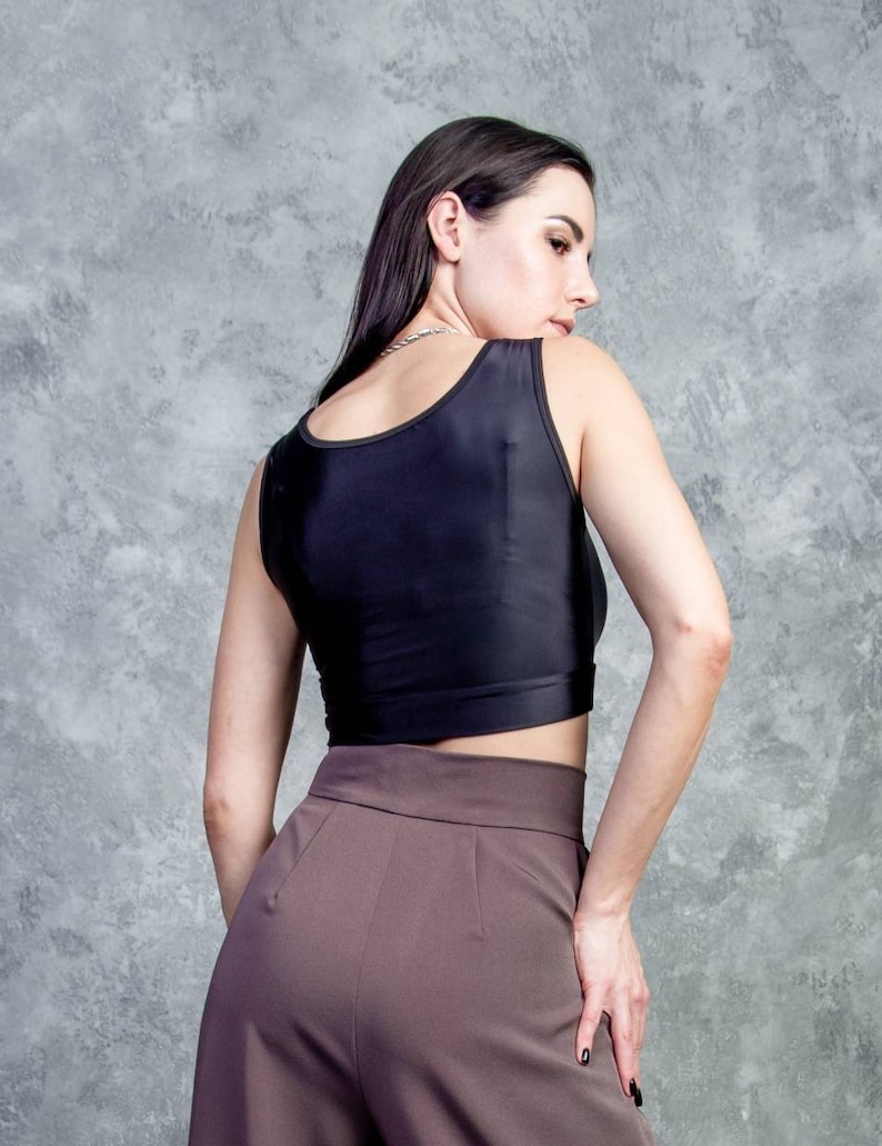 K&ouml;nnte beinhalten: Eine Frau tr&auml;gt ein schwarzes &auml;rmelloses Crop-Top und eine braune, hochgeschlossene Hose. Sie steht vor einer grauen, strukturierten Wand.