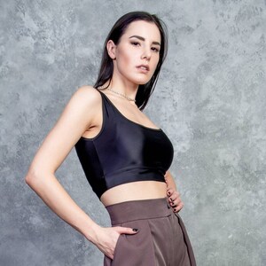 K&ouml;nnte beinhalten: Eine Frau tr&auml;gt ein schwarzes Crop-Top und eine braune, hochgeschlossene Hose. Sie steht vor einer grauen, strukturierten Wand.