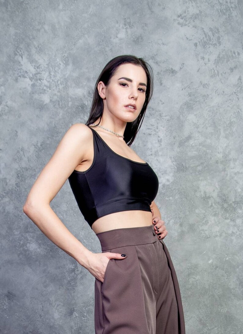 K&ouml;nnte beinhalten: Eine Frau tr&auml;gt ein schwarzes Crop-Top und eine braune, hochgeschlossene Hose. Sie steht vor einer grauen, strukturierten Wand.