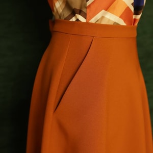 May include: A rust-coloured skirt with a single pocket on the front. The skirt is made of a smooth, woven fabric.